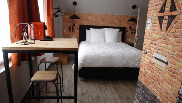 Ein Schlafzimmer mit Ziegelwand, einem Bett mit weißer Bettwäsche, einem Schreibtisch mit Stuhl und einer Pendelleuchte.