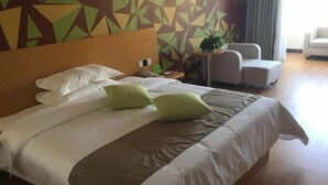 Quarto de hotel com uma cama grande, duas almofadas verdes, uma cabeceira de madeira e parede com padrão geométrico.