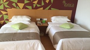 Quarto de hotel com duas camas, cada uma com travesseiro e cobertor. Tem uma mesa de cabeceira com telefone e uma pequena mesa com abajur. O quarto tem parede com padrão geométrico e um letreiro que diz "Vatica".