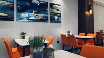 A dining area with orange chairs and tables, a potted plant, and wall art.