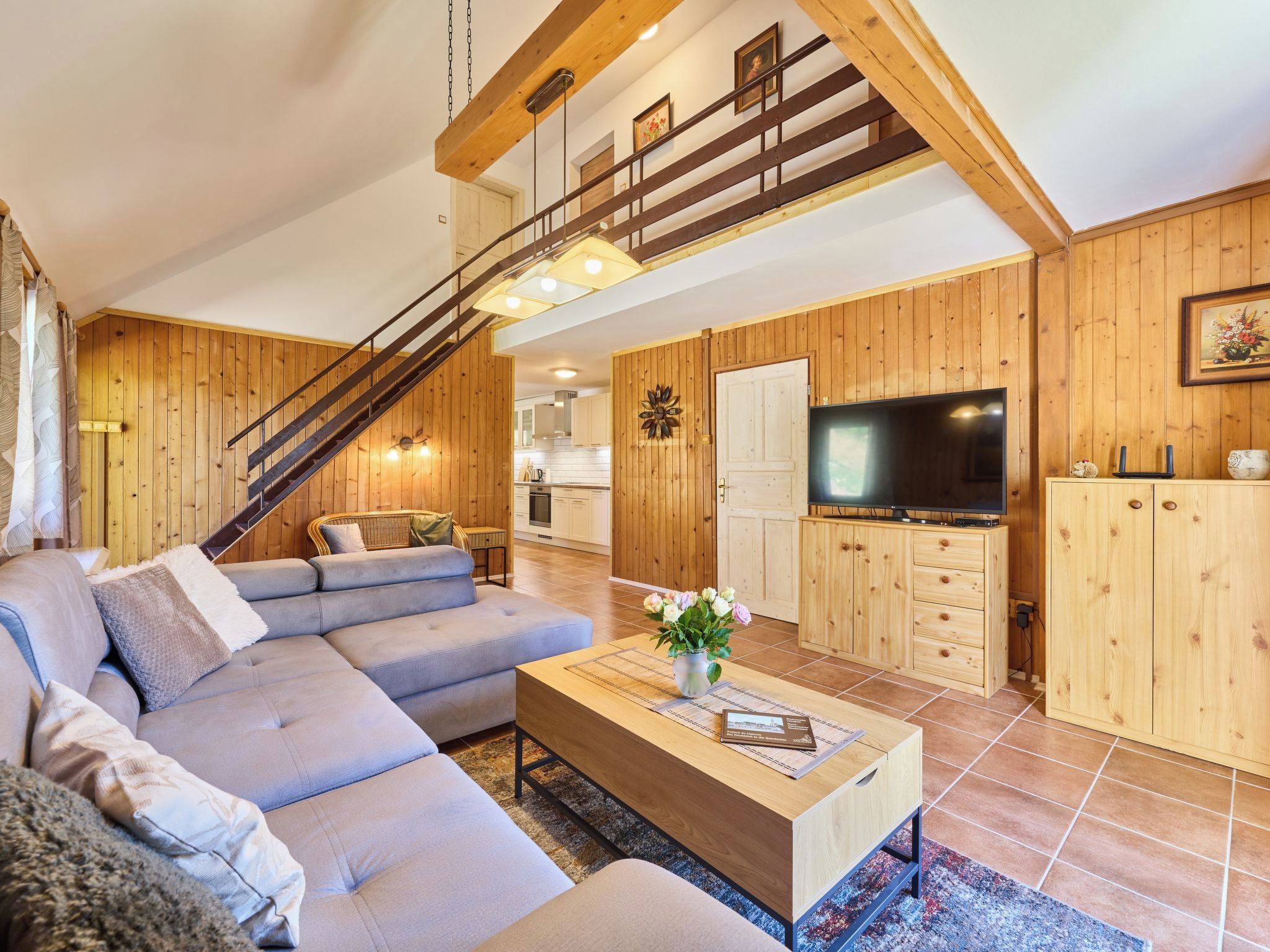 Un salon moderne avec un canapé d’angle, un escalier en bois, un téléviseur à écran plat et une cuisine en arrière-plan.