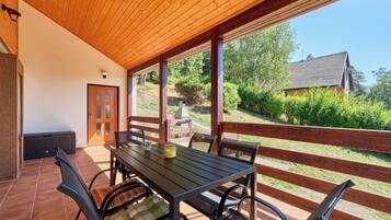 Une terrasse couverte avec une table et des chaises, une porte et une vue sur une maison et des arbres.