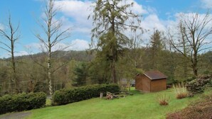Een klein houten huisje, omringd door grasvelden, bomen en een heldere lucht.