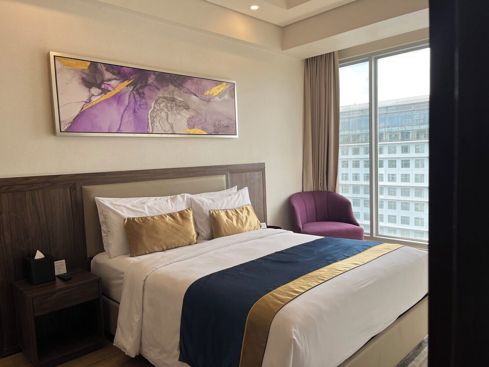A hotel room with a large bed, a nightstand, a chair, and a window with a city view.