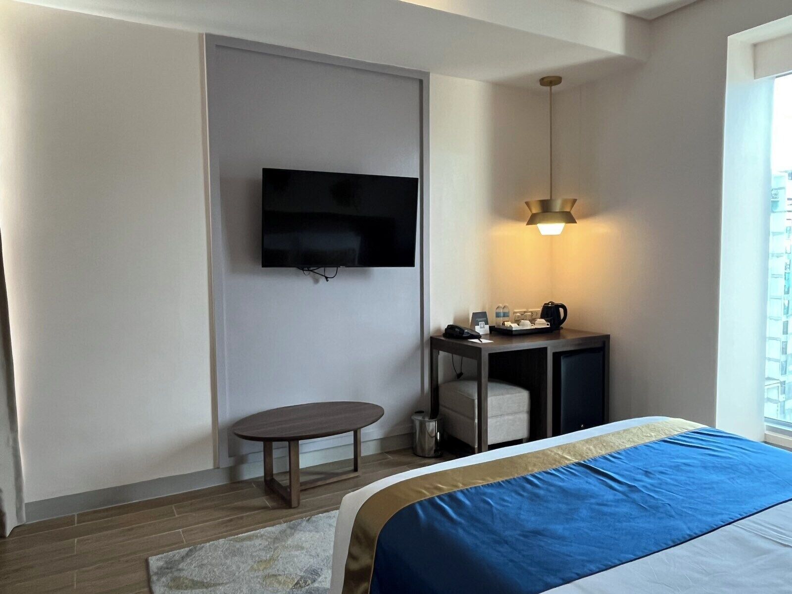 Quarto de hotel com cama, televisão fixada na parede, mesinha com abajur e janela com vista.