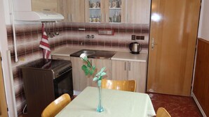 Een kleine keuken met een fornuis, spoelbak en kastjes. Er staat een eettafel met stoelen en een vaas met een bloem.