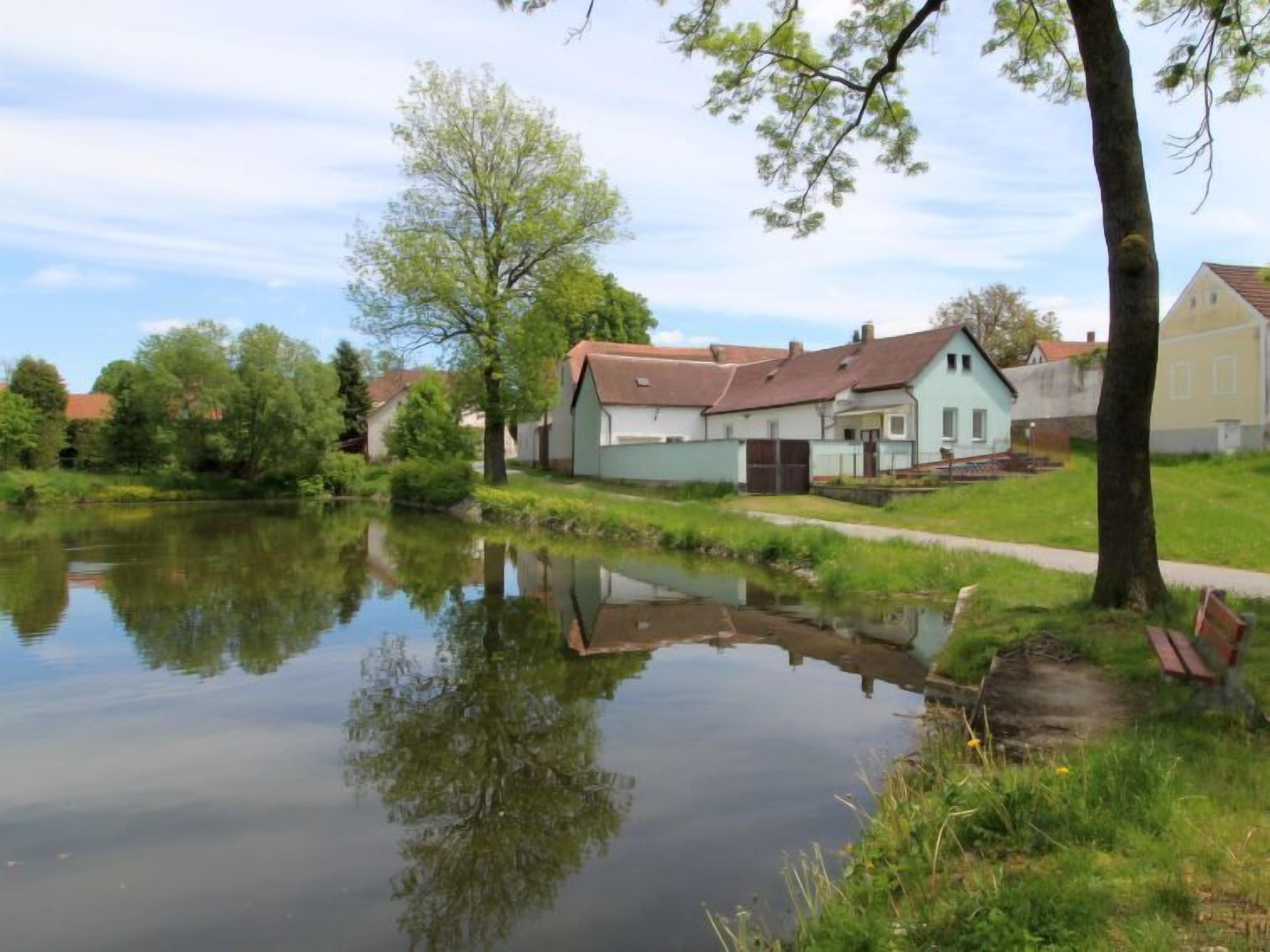 Ein ruhiger Fluss spiegelt Gebäude und Bäume wider, mit einem Pfad, der zu einem hellen Haus mit rotem Dach führt.