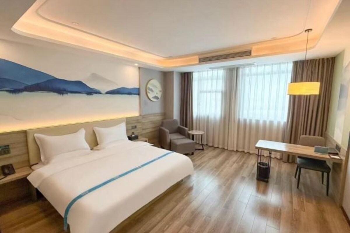 A hotel room with a large bed, a desk, a chair, a small table, a lamp, and a painting on the wall.