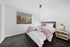Une chambre moderne avec une grande fenêtre, un lit recouvert d’une couverture blanche et rose, une table de chevet avec une lampe et une petite étagère en bois.