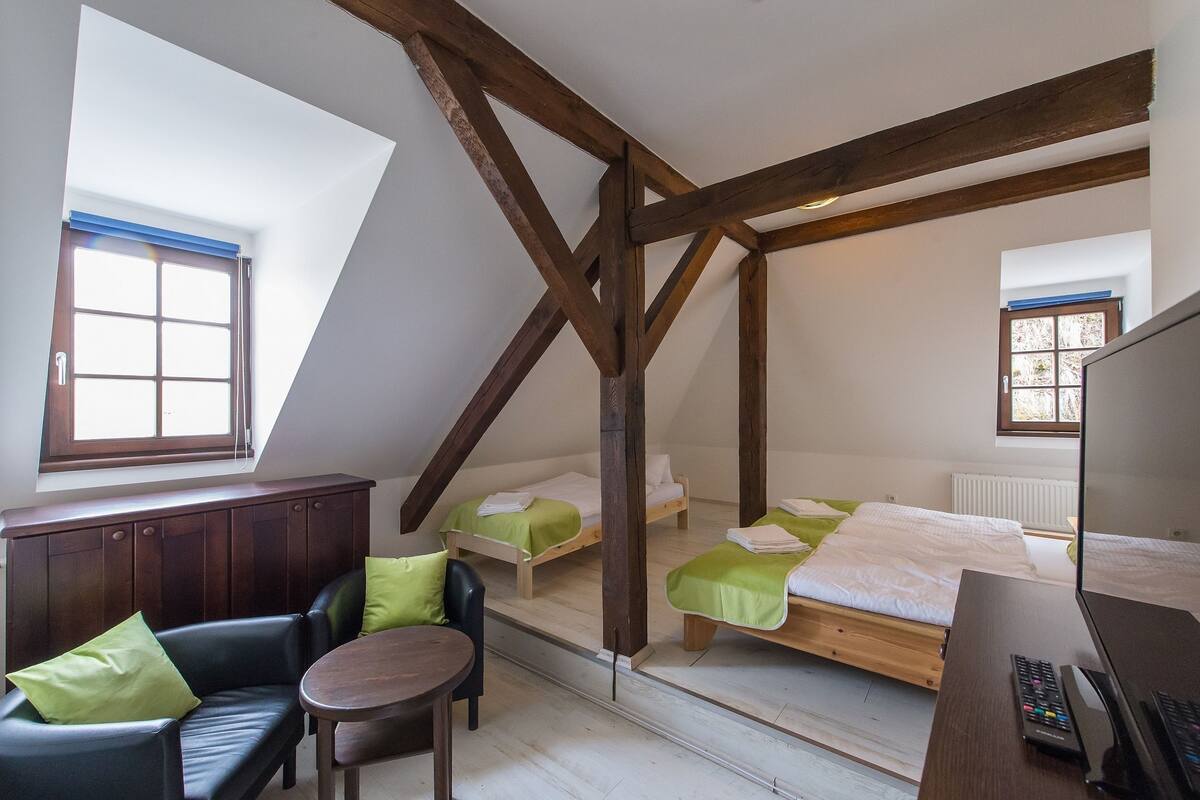 A room with a sloped ceiling, wooden beams, a bed, a desk, a chair, and a window.
