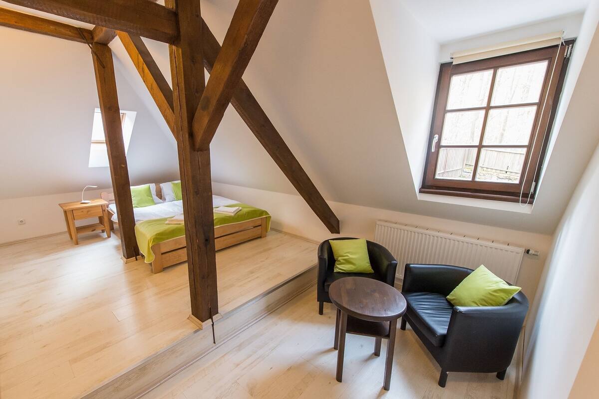 A loft-style room with a sloped ceiling, a bed, a sofa, a small table, and a window with a view.