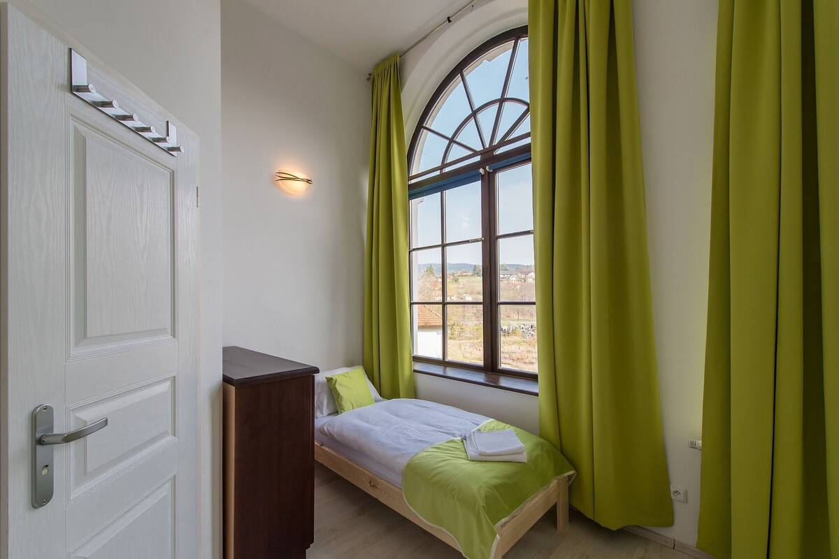 A bedroom with a bed, a window with curtains, a door, and a wall-mounted light.
