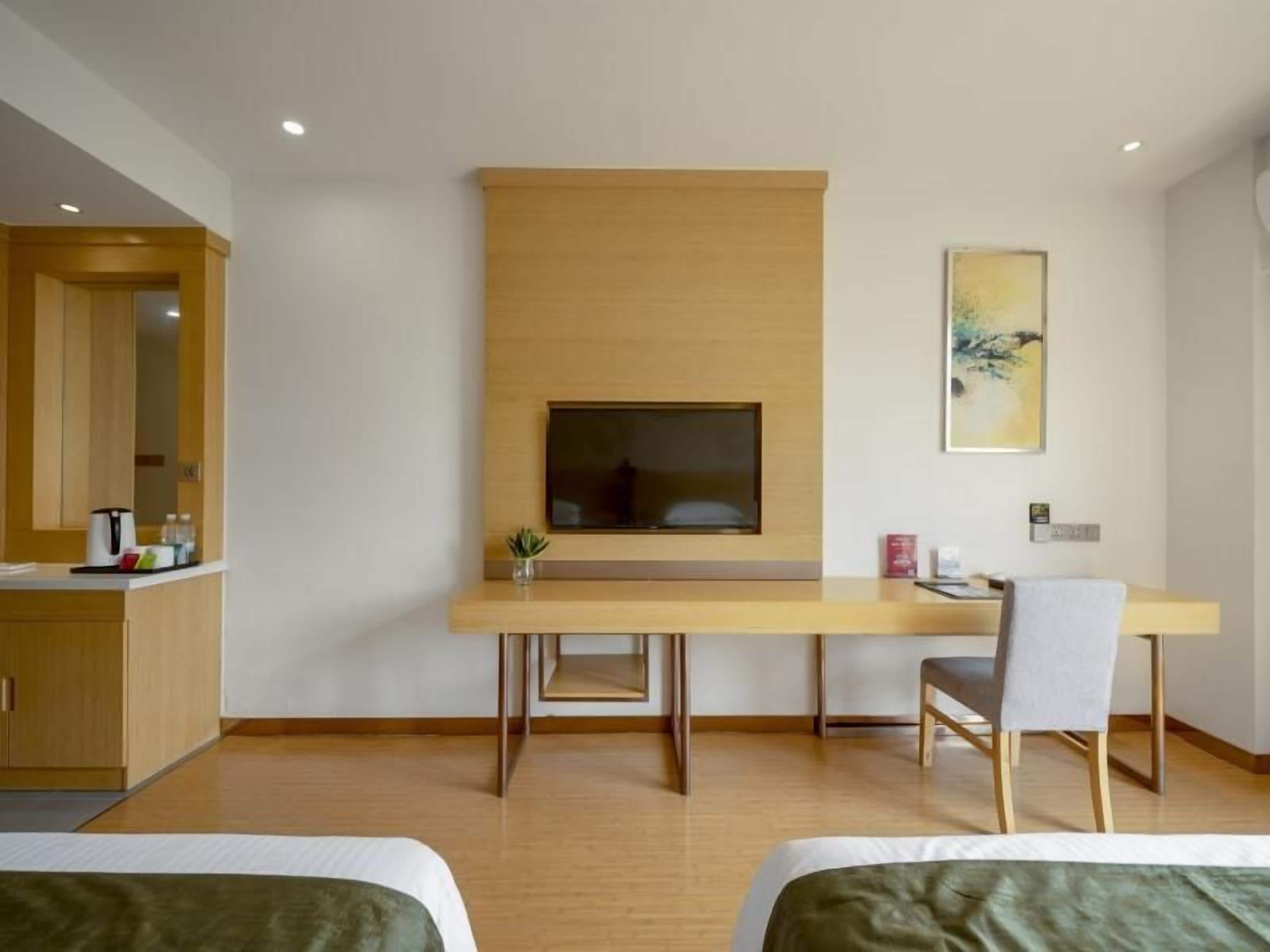 A modern hotel room with a wooden desk, a television mounted on the wall, a bed with green bedding, and a small potted plant.