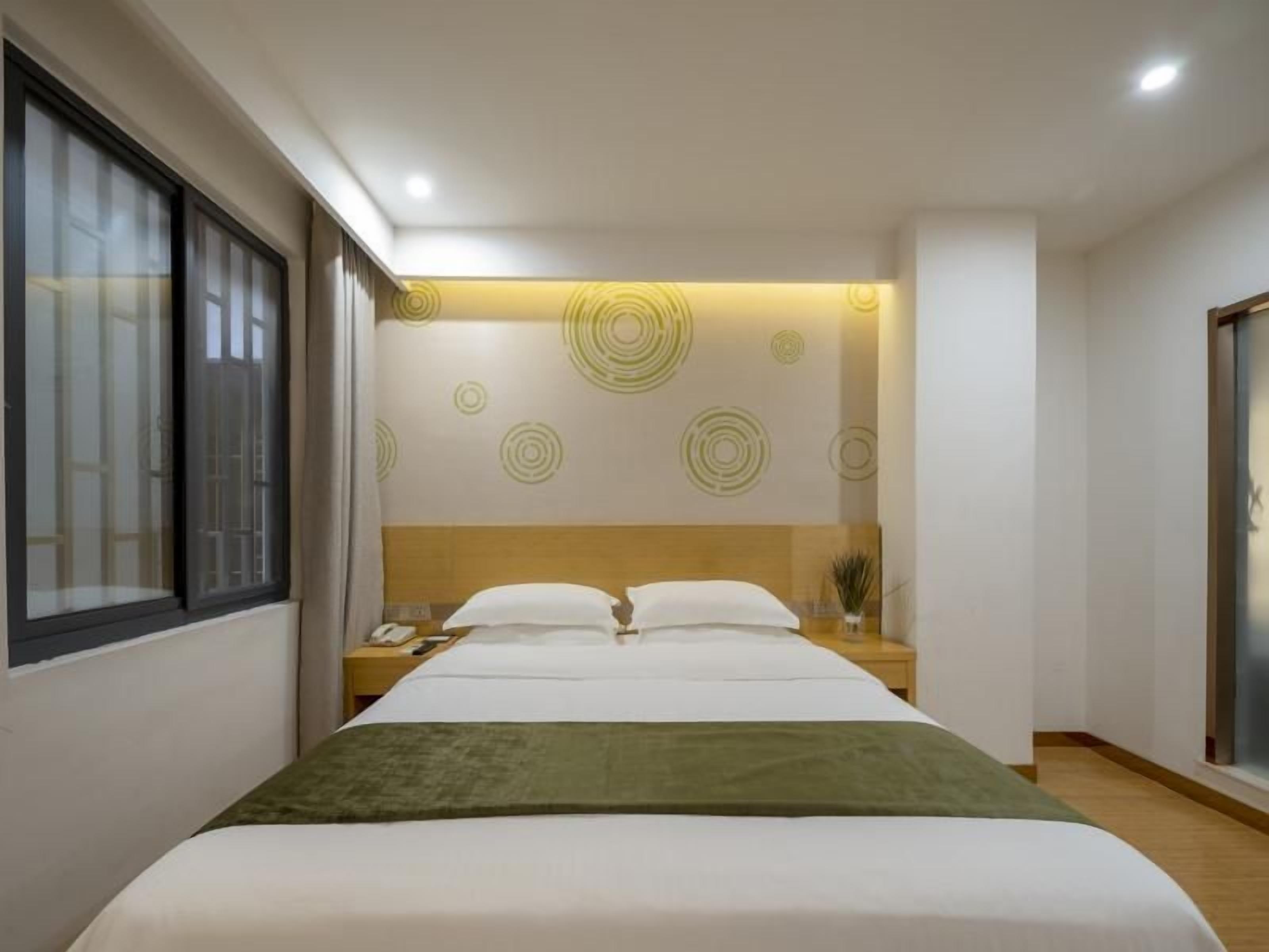 A modern hotel room with a large bed, a bedside table with a lamp, and a window with blinds.