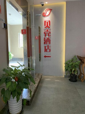 A hotel reception area with a modern design, featuring a reception desk, a security camera, and potted plants. - Shell Shanghai Chedun Yingshi City Yingshi Road Hotel (Shanghai)