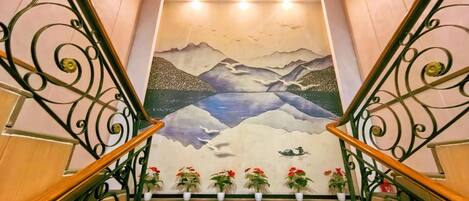 A staircase with a mural of mountains and water, flanked by ornate metal railings and potted plants.