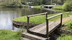 Un muelle de madera con barandillas se extiende sobre un lago tranquilo, rodeado de exuberante vegetación y un puente al fondo.