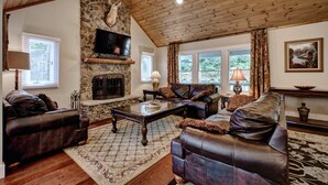 Una sala de estar acogedora con sofás de cuero, chimenea de piedra, televisor y ventanas amplias.