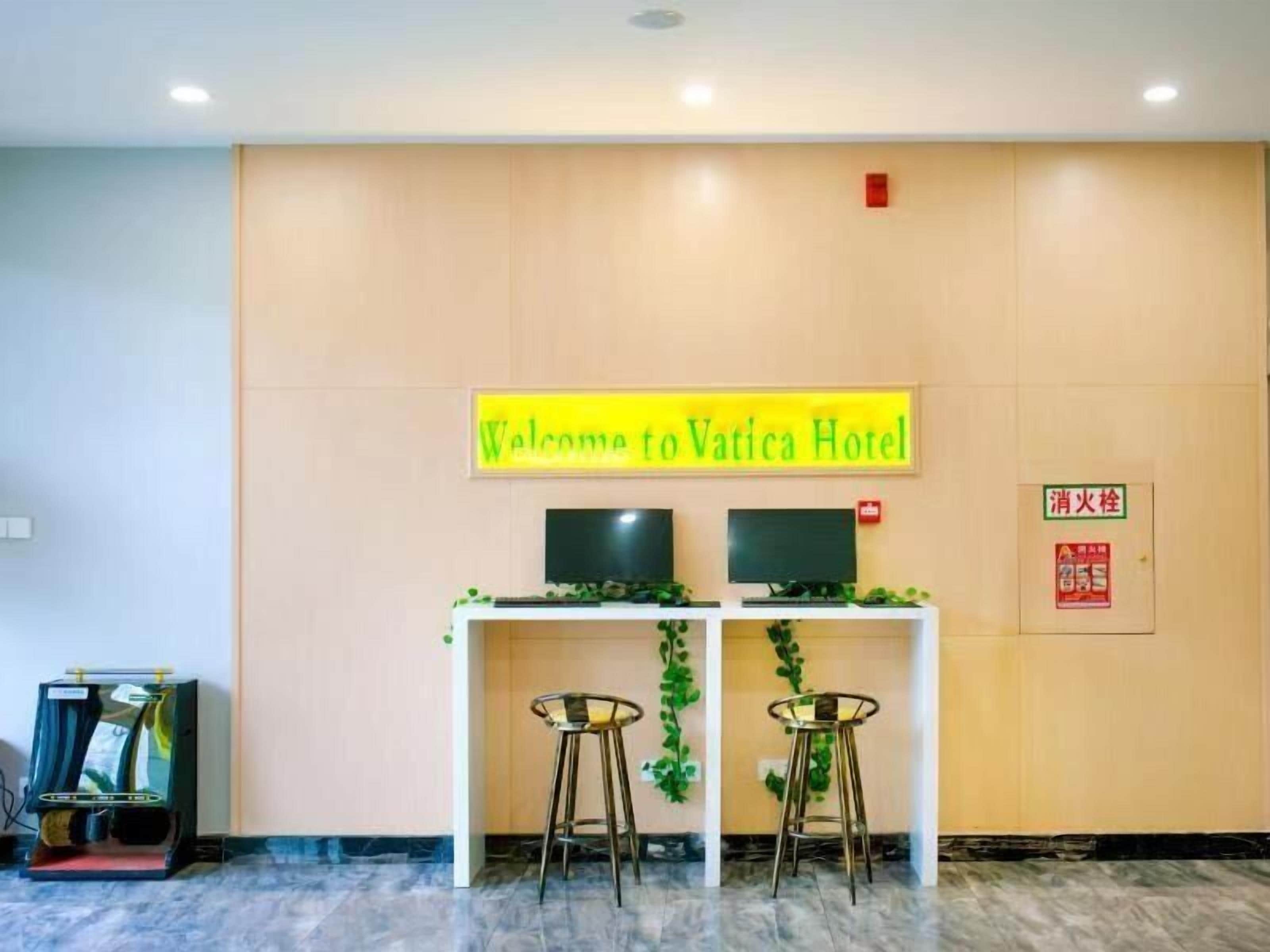 Una recepción con un cartel que dice 'Welcome to Vatica Hotel', dos monitores y dos taburetes de bar.