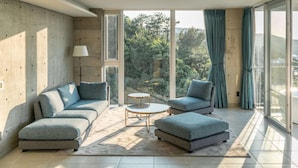 A modern living room with a sofa set, a round coffee table, and a view of a forest through large windows. - Okinawa Plaisant Villa (Onna)