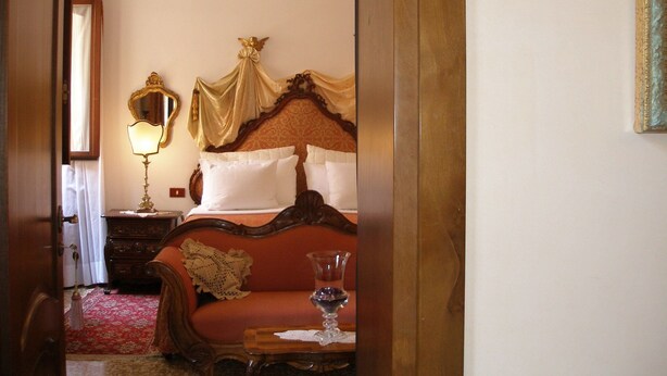 A hotel room with a bed, a bedside table with a lamp, a chandelier, and a red carpet.