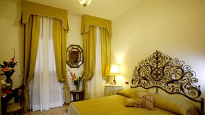 A hotel room with a bed, a nightstand with a lamp, a mirror, curtains, and a floral arrangement.