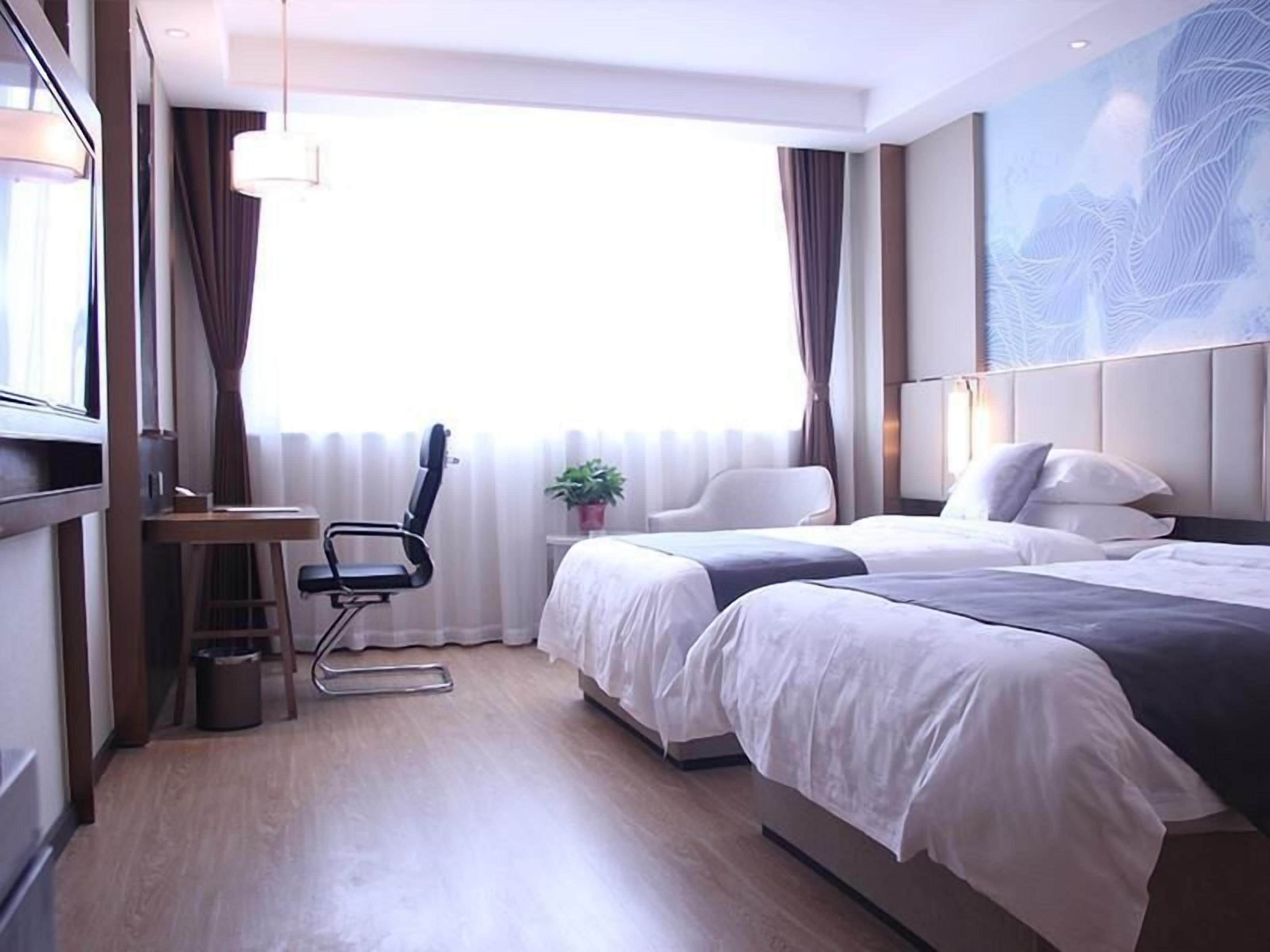 A hotel room with two beds, a desk, a chair, a potted plant, and a large window with curtains.