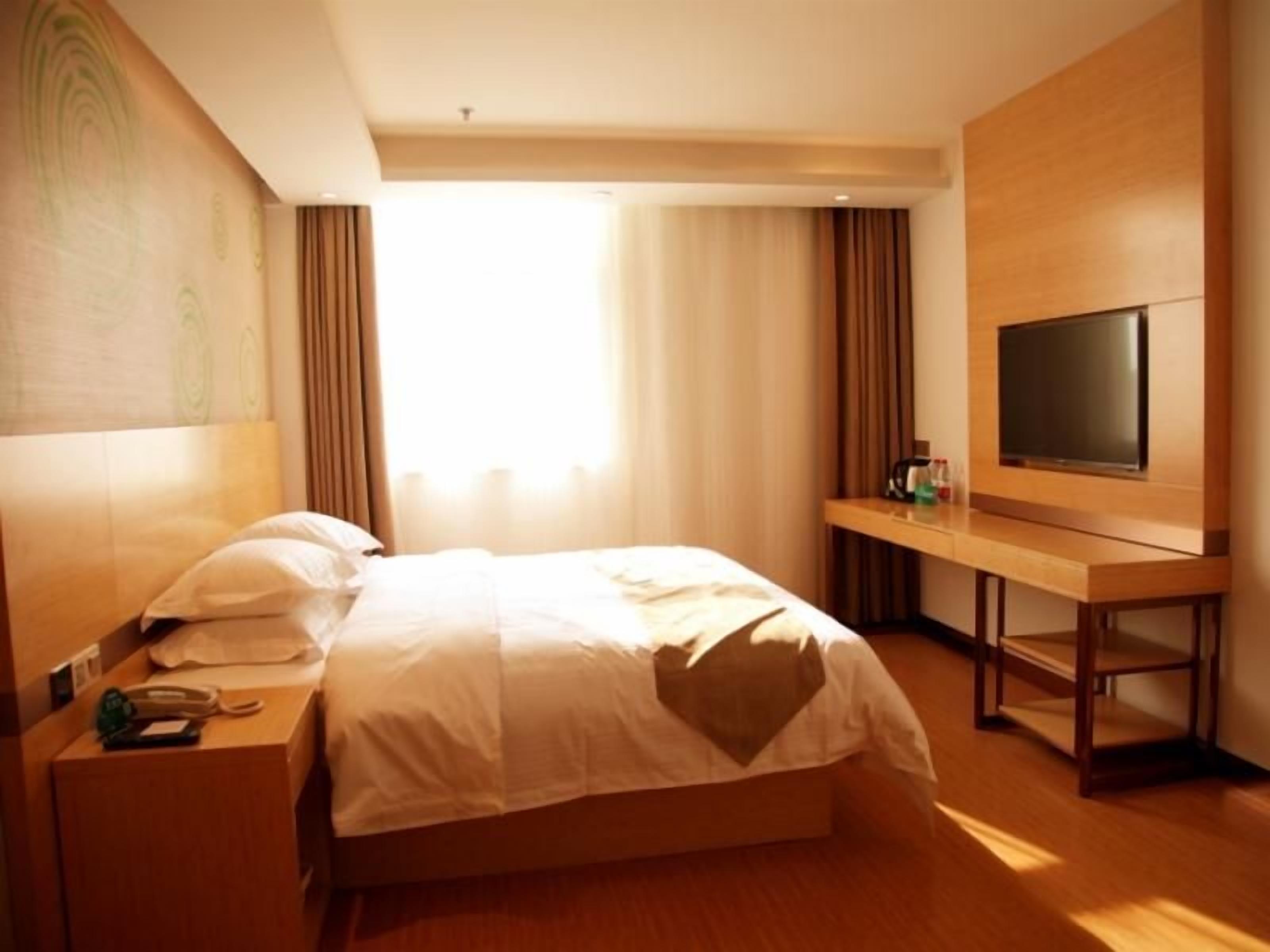 A hotel room with a bed, a bedside table with a phone and a bottle, a TV on a wooden stand, and a window with curtains.