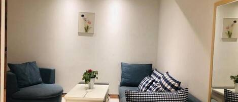 Una sala de estar moderna con un sofá azul oscuro, un sillón más pequeño y una mesita de noche blanca con un jarrón de flores.