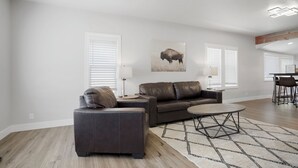 A modern living room with a brown leather sofa, a matching armchair, and a coffee table. There is a painting of a bison on the wall, and the room has large windows with blinds. - Newly Re-Finished 2 bed, 1 bath close to downtown (Sheridan)