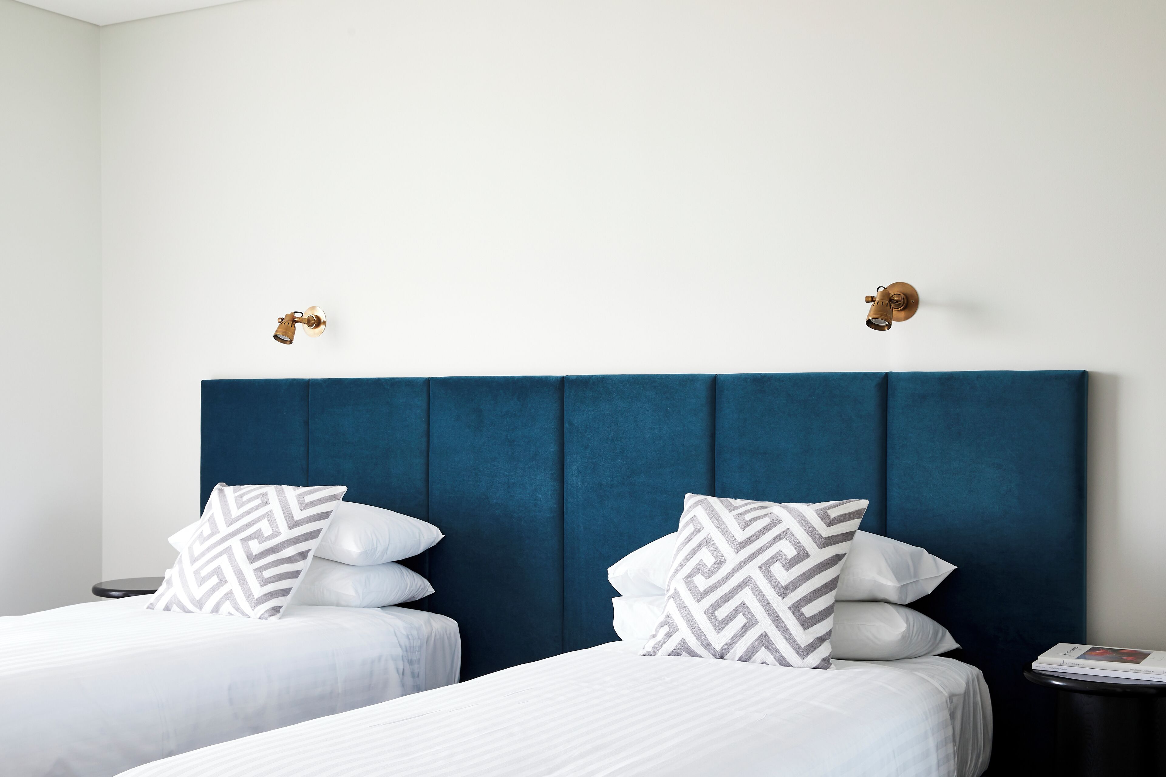 A hotel room with two beds, each with white bedding and patterned pillows, a dark blue headboard, and a small side table with a book.