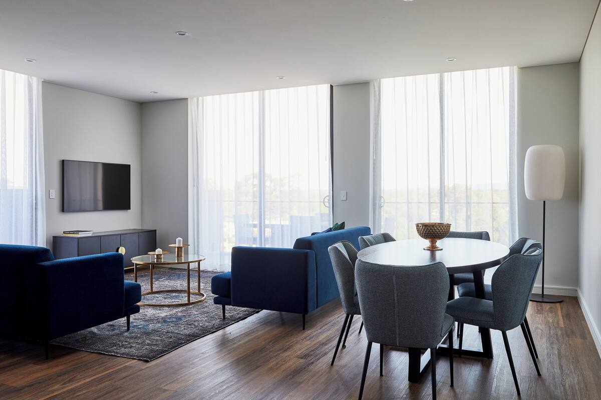 A modern living room with a sofa, a dining table with chairs, a flat-screen TV mounted on the wall, and large windows with sheer curtains.