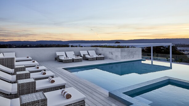 A rooftop infinity pool with white lounge chairs and a view of the cityscape at sunset.
