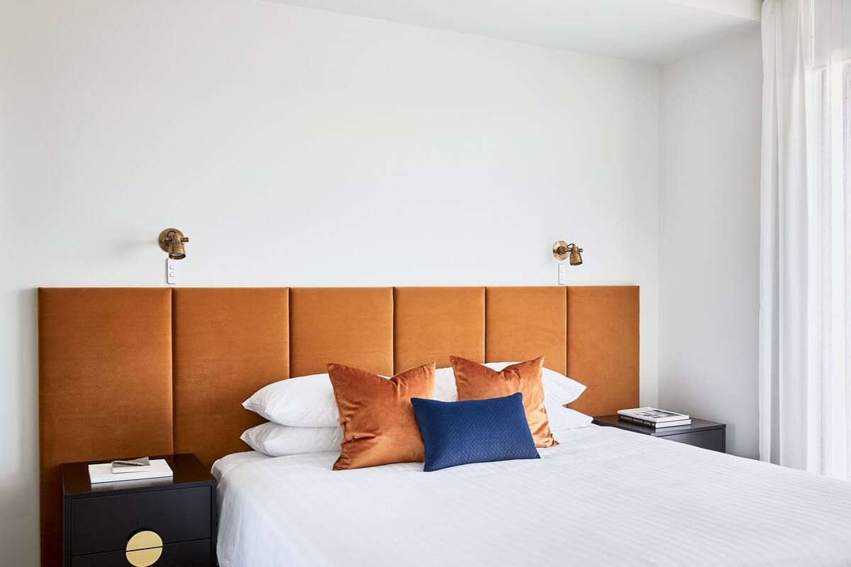 A neatly made bed with a brown headboard, white pillows, and a blue accent pillow. There is a nightstand with a book and a lamp.