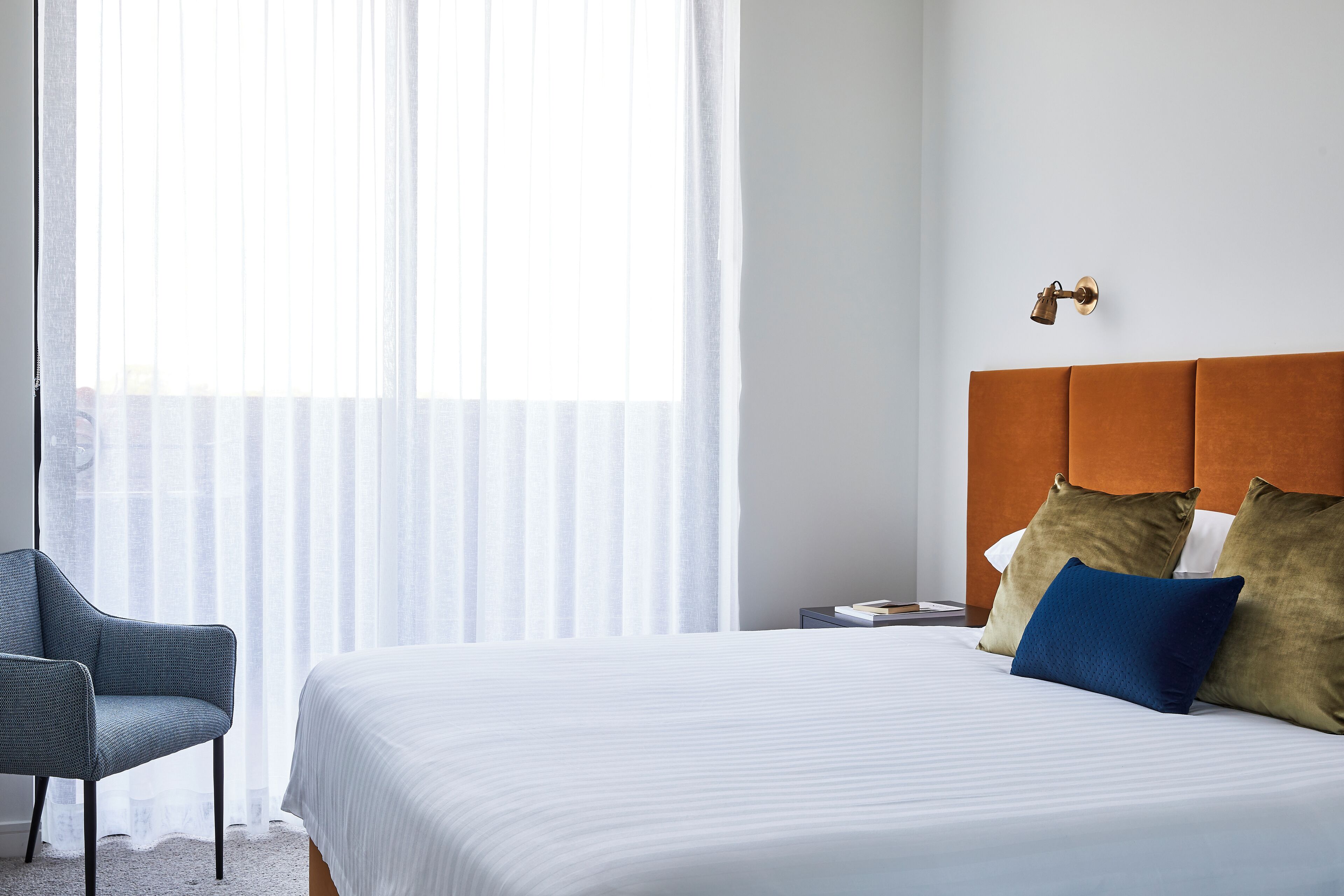 A hotel room with a bed, a chair, a nightstand, and a window with sheer curtains.
