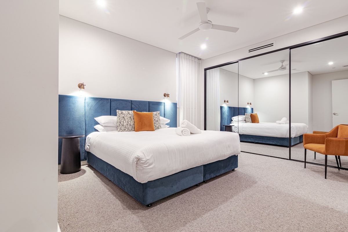 A modern hotel room with a large bed, a small table, a chair, and a large mirror.