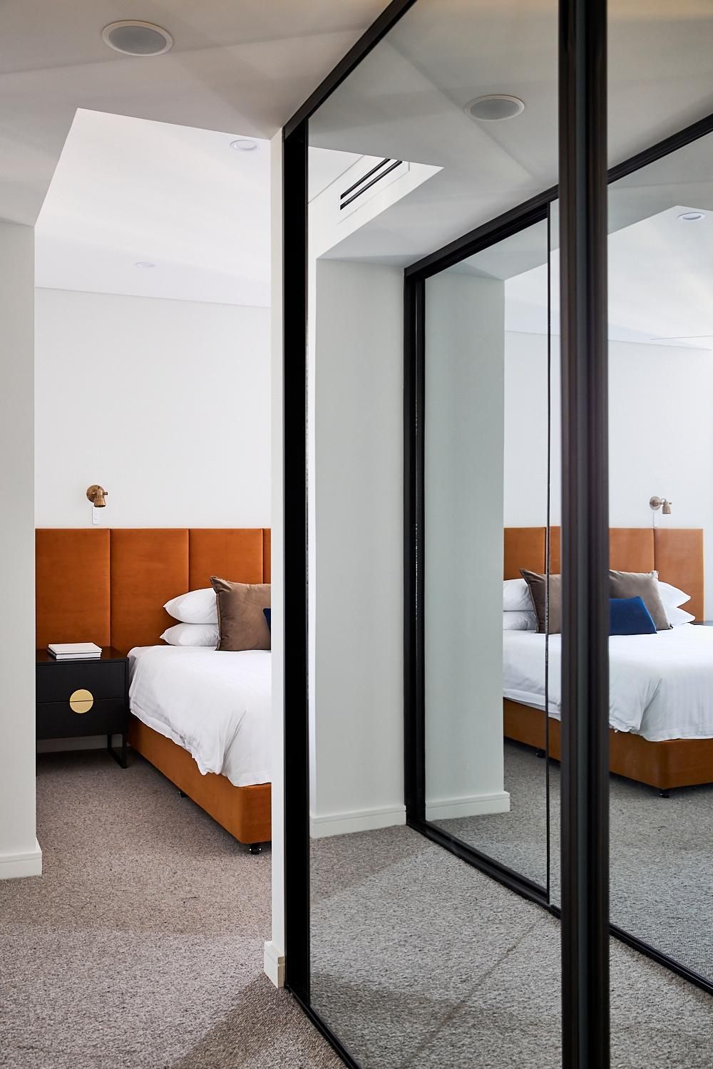 A modern hotel room with a large mirror, a bed with white bedding, a nightstand with a book, and a wall-mounted light fixture.
