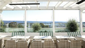 A sunlit outdoor seating area with tables and wicker chairs, overlooking a cityscape and hills.