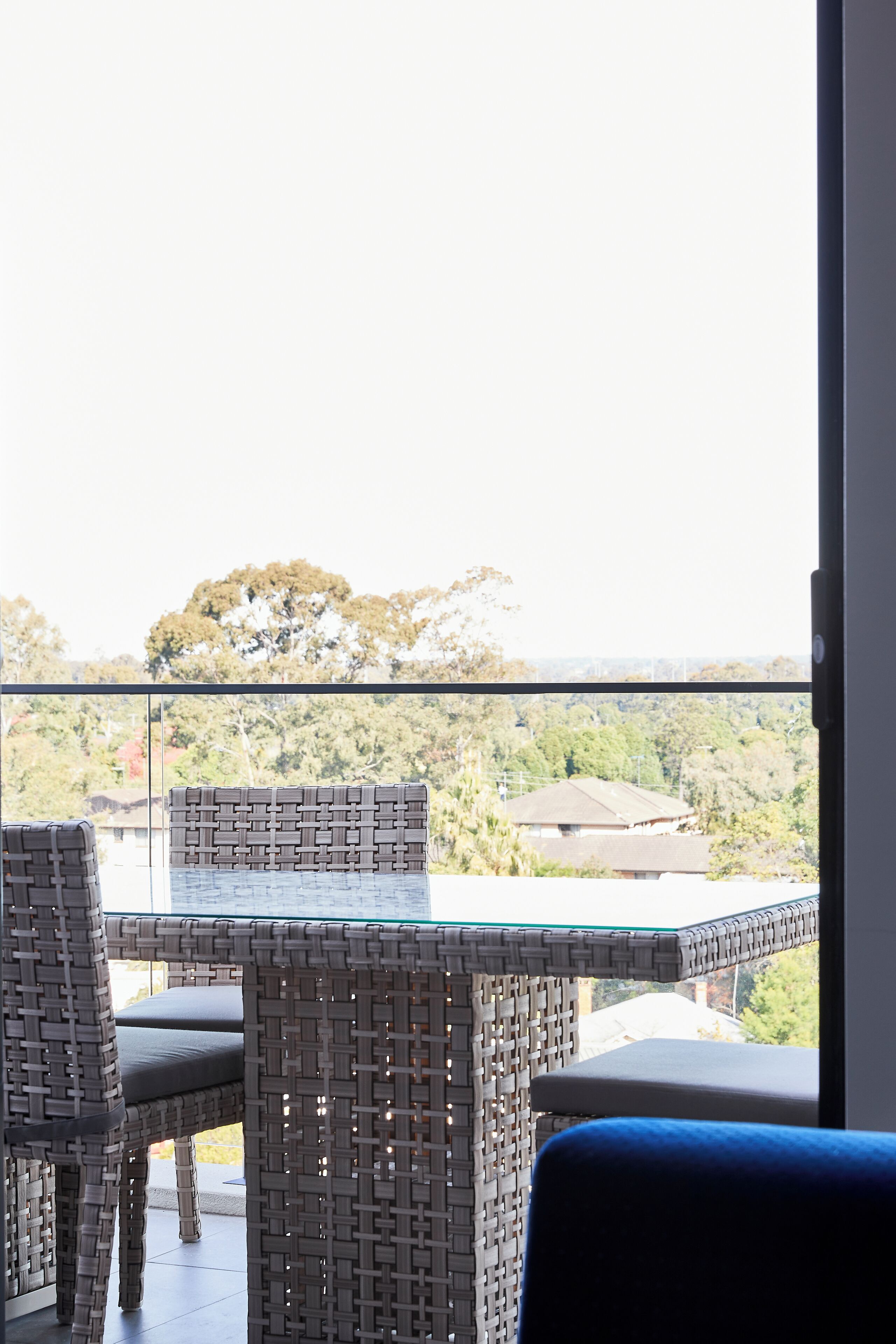 A balcony with wicker furniture and a glass table, overlooking a scenic landscape with trees and buildings.