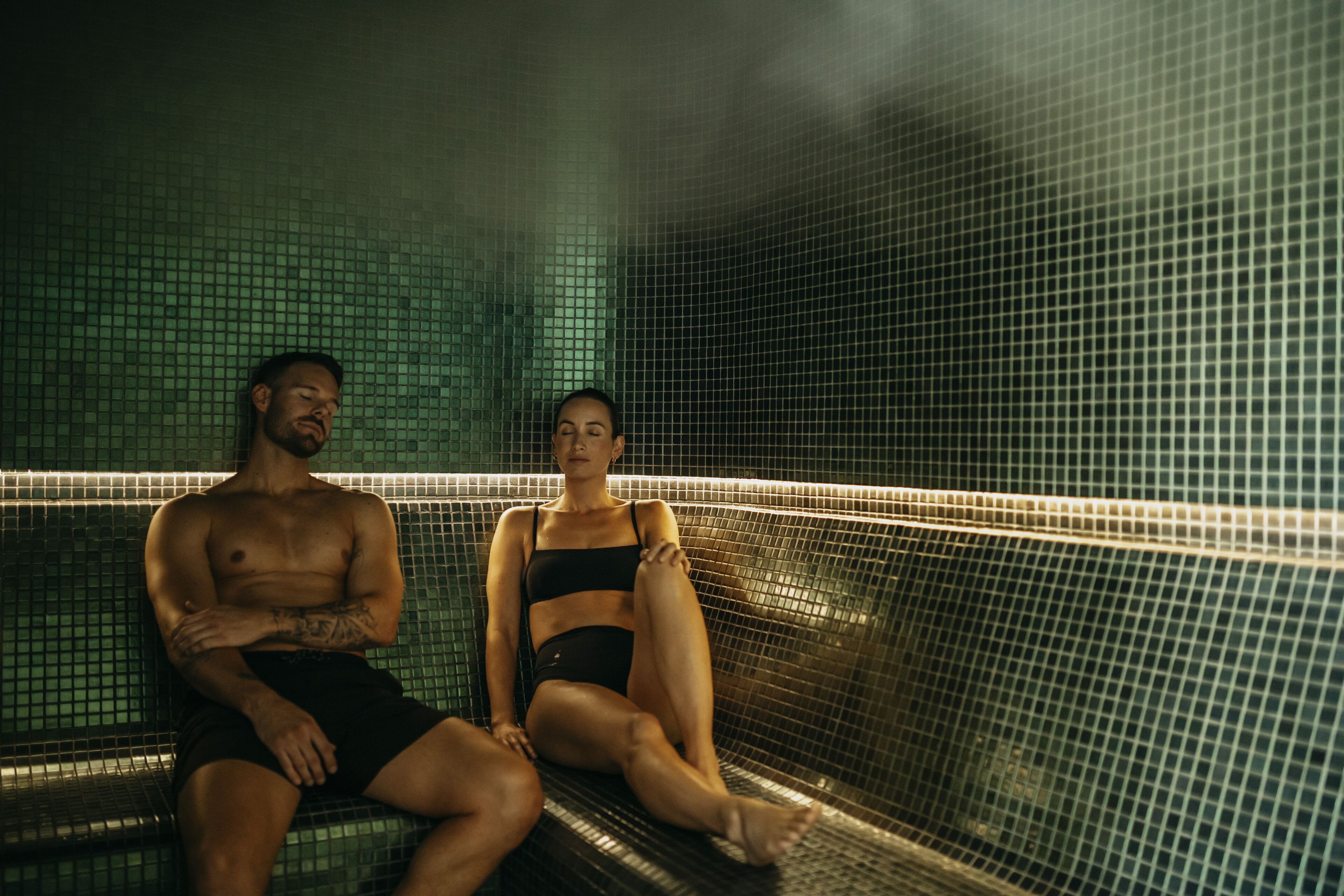 Two people sitting on a bench in a tiled room.