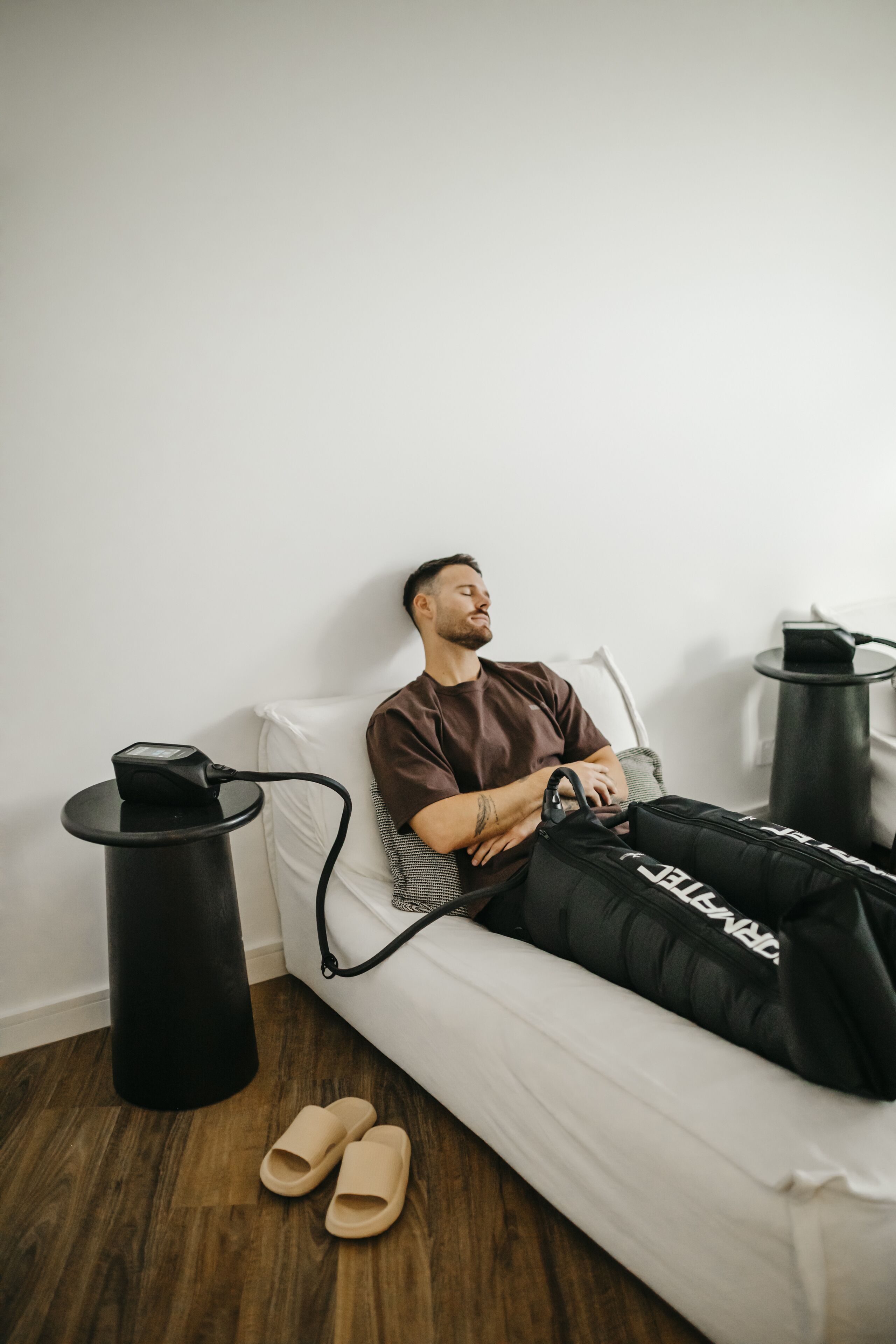 A person lying on a white couch with a black bag beside them, a black trash bin, and a pair of beige slippers on the floor.