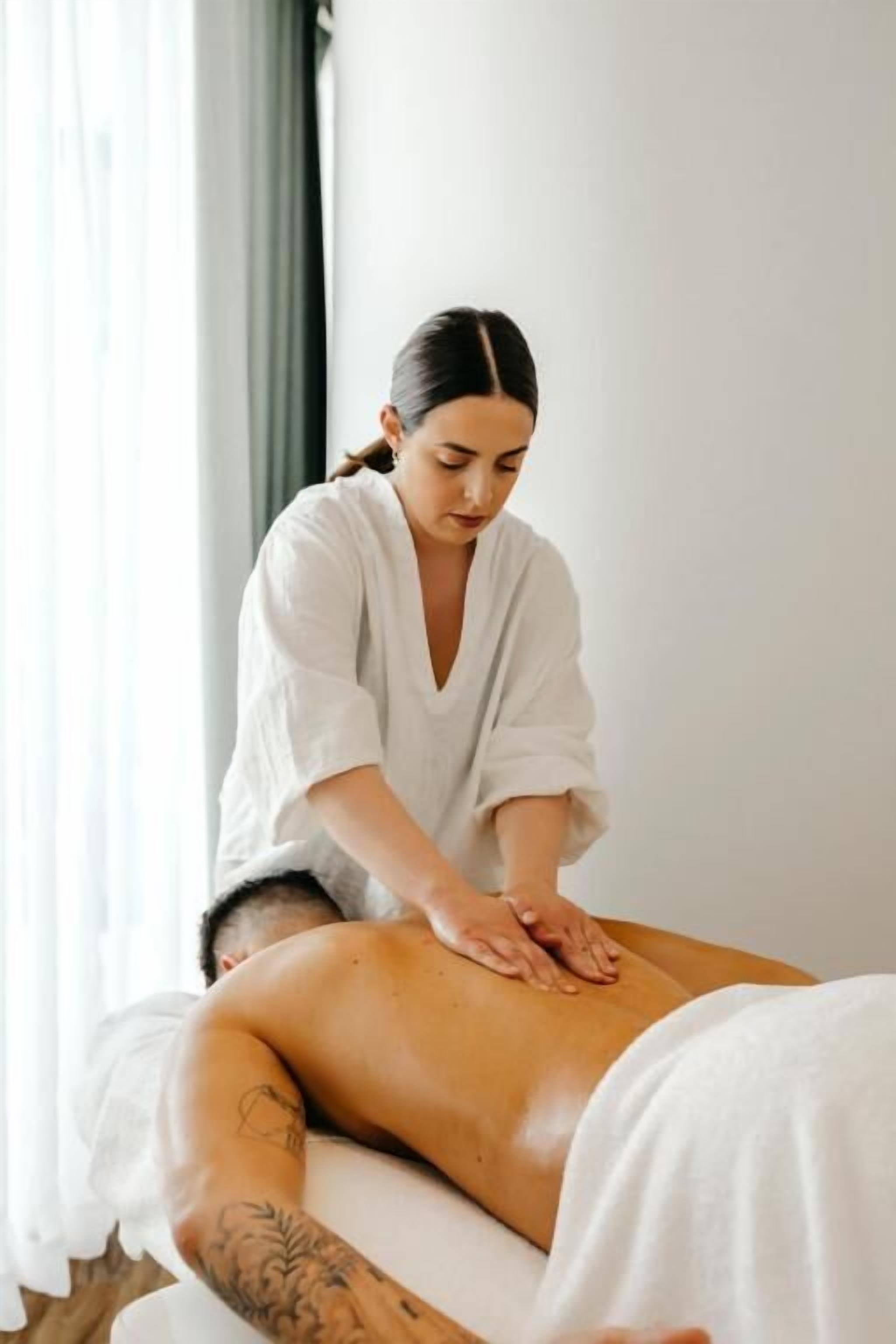 A massage therapy session in a room with a bed and a window with curtains.