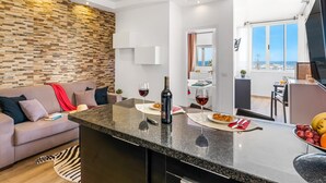 A modern living room with a stone wall, a sofa, a dining area with a table, and a kitchen visible in the background. - Rosamar 313 Luxury sea view wifi (Arona)