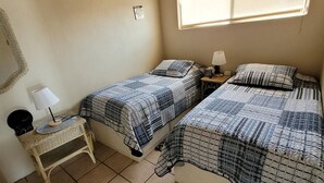 A bedroom with two beds, a small bedside table with a lamp, a wicker chair, and a window providing natural light. - CASA MARBELLA 2BR/1BA Private Beach-Front Home (Ensenada)