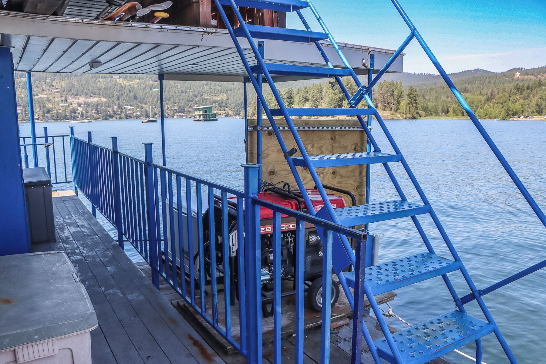 Una escalera metálica azul que conduce a una terraza con un motor de barco y una estructura de madera al fondo.