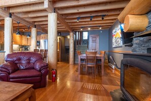 Interior em madeira com sofá de couro, mesa de jantar e cadeiras. Uma lareira é visível à direita.