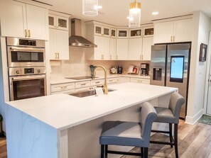 Cocina moderna con armarios blancos, electrodomésticos de acero inoxidable, una isla grande con taburetes y luminarias colgantes.