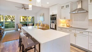 A modern kitchen with white cabinets, a large island, and a dining area with a view of the outdoors.