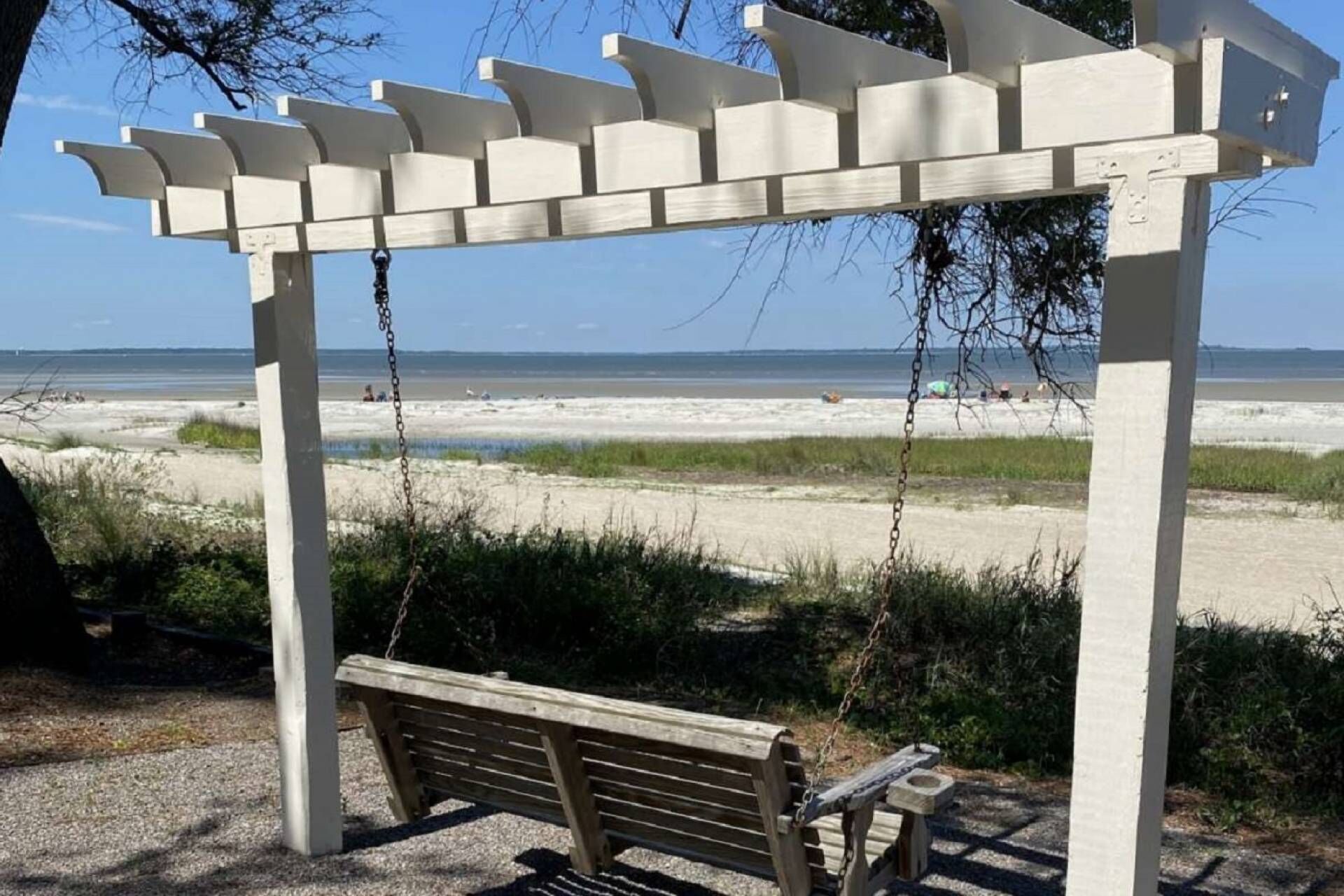 A wooden bench under a white pergola with a view of the beach and ocean.