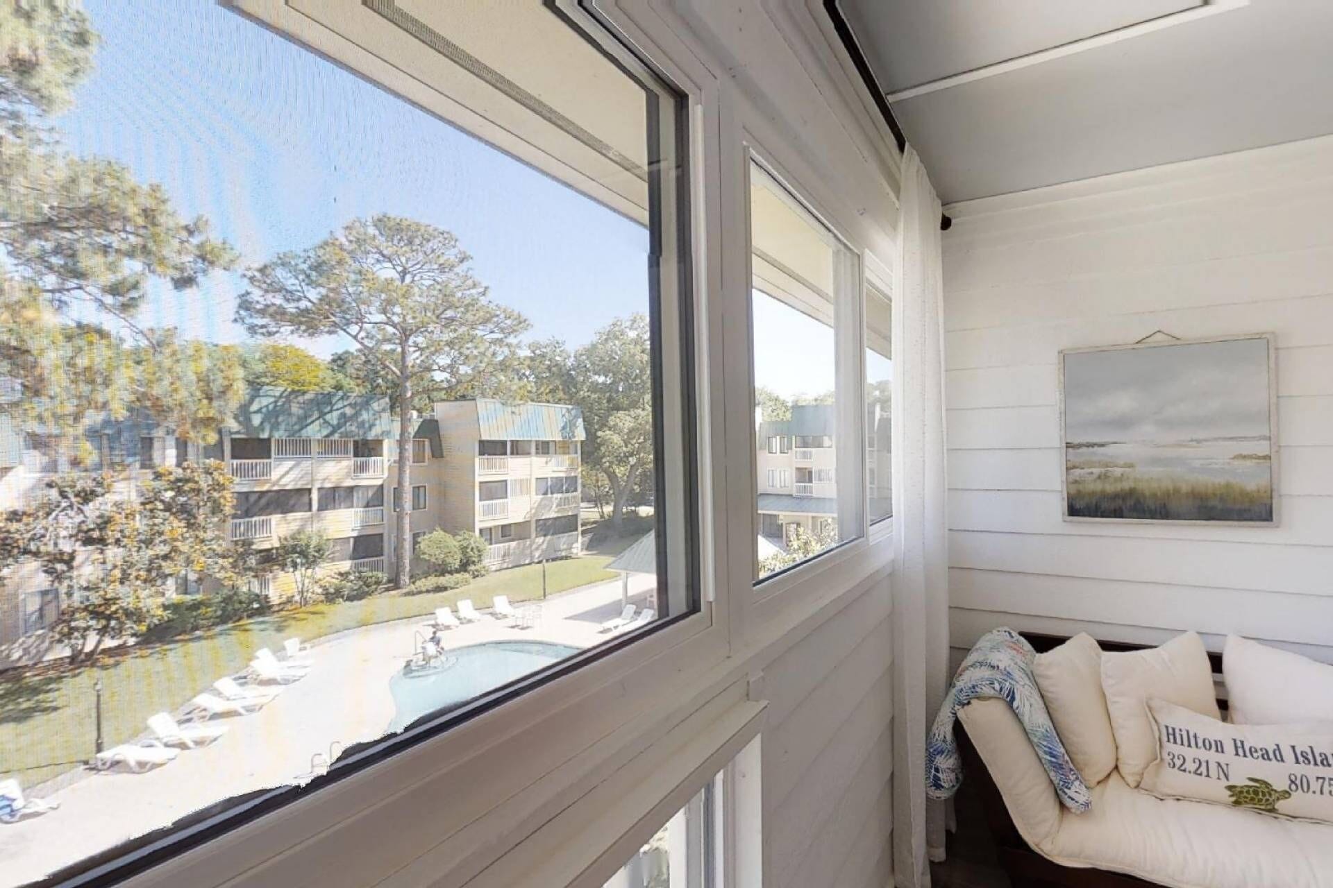 Ein Zimmer mit Blick auf einen Pool und ein Gebäude gegenüber. Ein Sessel mit einem Kissen mit der Aufschrift 'Hilton Head Island 32.21N 80.75'.
